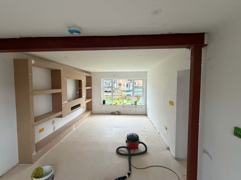 An unfinished lounge with bare plaster walls, raw MDF shelving, a red steel beam and dust sheets